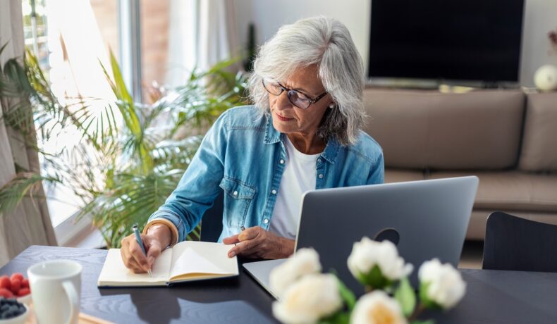 Femeie în vârstă, cu părul alb și cămașă albastră, care stă la birou, în fața unui laptop, care scrie într-un jurnal, pentru a ilustra o „mică bunicuță” care riscă 10 ani de închisoare